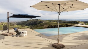 Un espace aménagé au bord de la piscine, avec vue sur des collines verdoyantes, comprenant un fauteuil de relaxation, une table et un parasol.