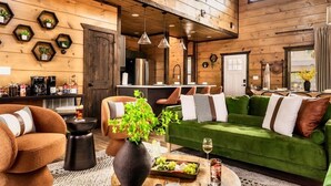 Un salon chaleureux avec un canapé vert, un fauteuil marron et une table basse sur laquelle sont disposées des boissons et des en-cas.