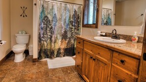Une salle de bain comprenant des toilettes, un lavabo, un meuble-lavabo en bois, un miroir et un rideau de douche orné d’un motif de forêt.