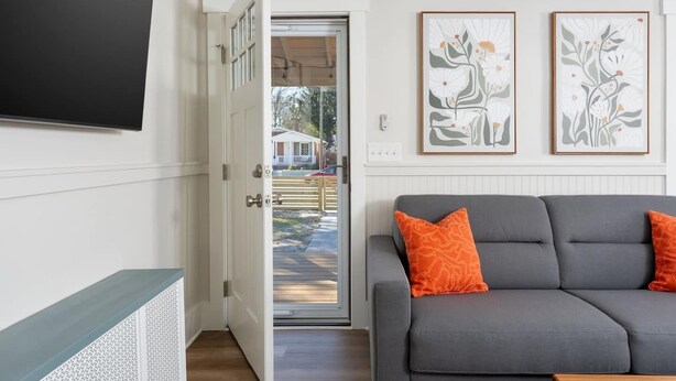 Ein Wohnzimmer mit grauem Sofa, einer offenen Tür, die nach draußen führt, und zwei gerahmten Blumenbildern an der Wand.