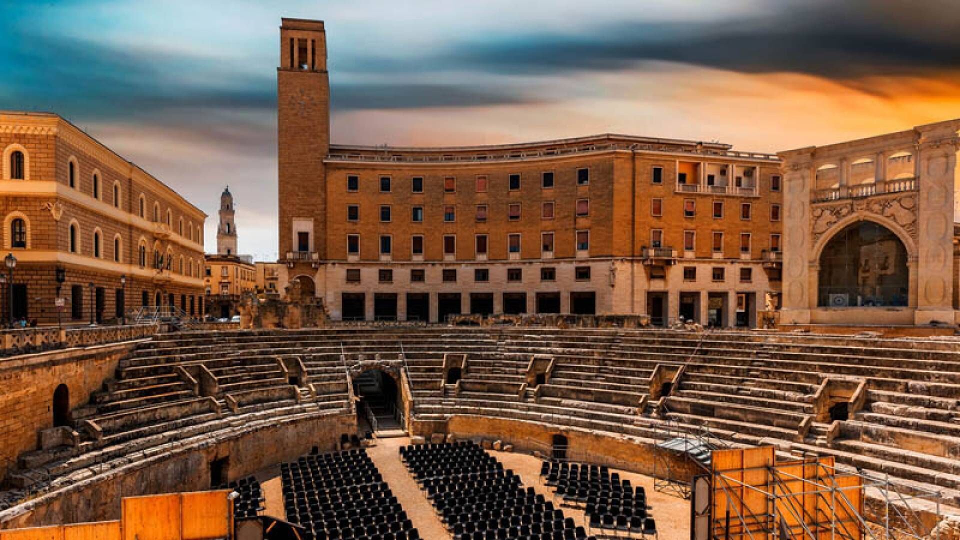 An ancient amphitheater with stone steps and a central stage, surrounded by classical architecture.