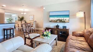Una sala de estar con un sofá color beige, un sofá de cuero marrón, un comedor con sillas, una mesa de centro con flores, un televisor mostrando una escena de playa y una lámpara.