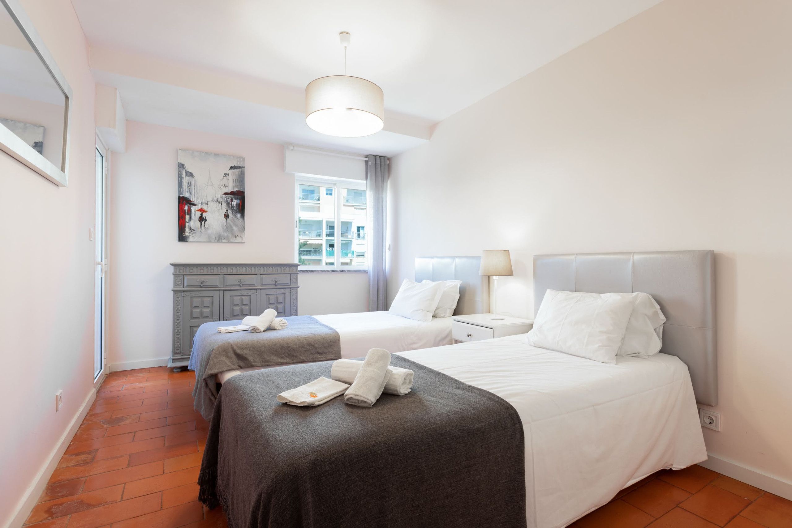 A hotel room with two beds, a dresser, a painting, and a window with curtains.