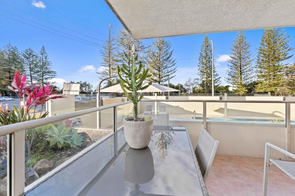 A balcony with a glass table, a potted plant, and a view of trees and a building.