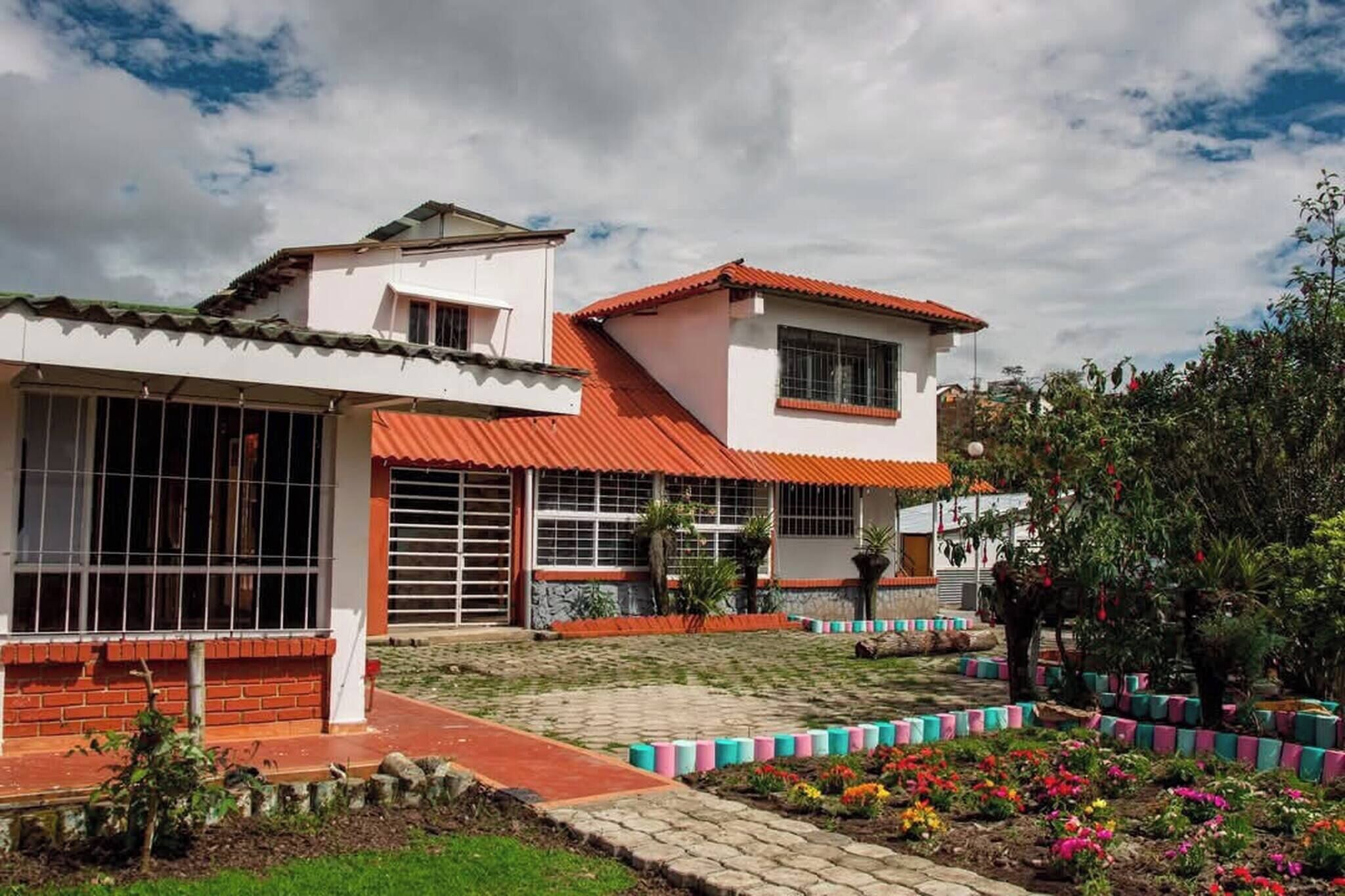 Un edificio de dos plantas con tejado de tejas rojas, amplias ventanas y un patio empedrado con coloridos parterres.
