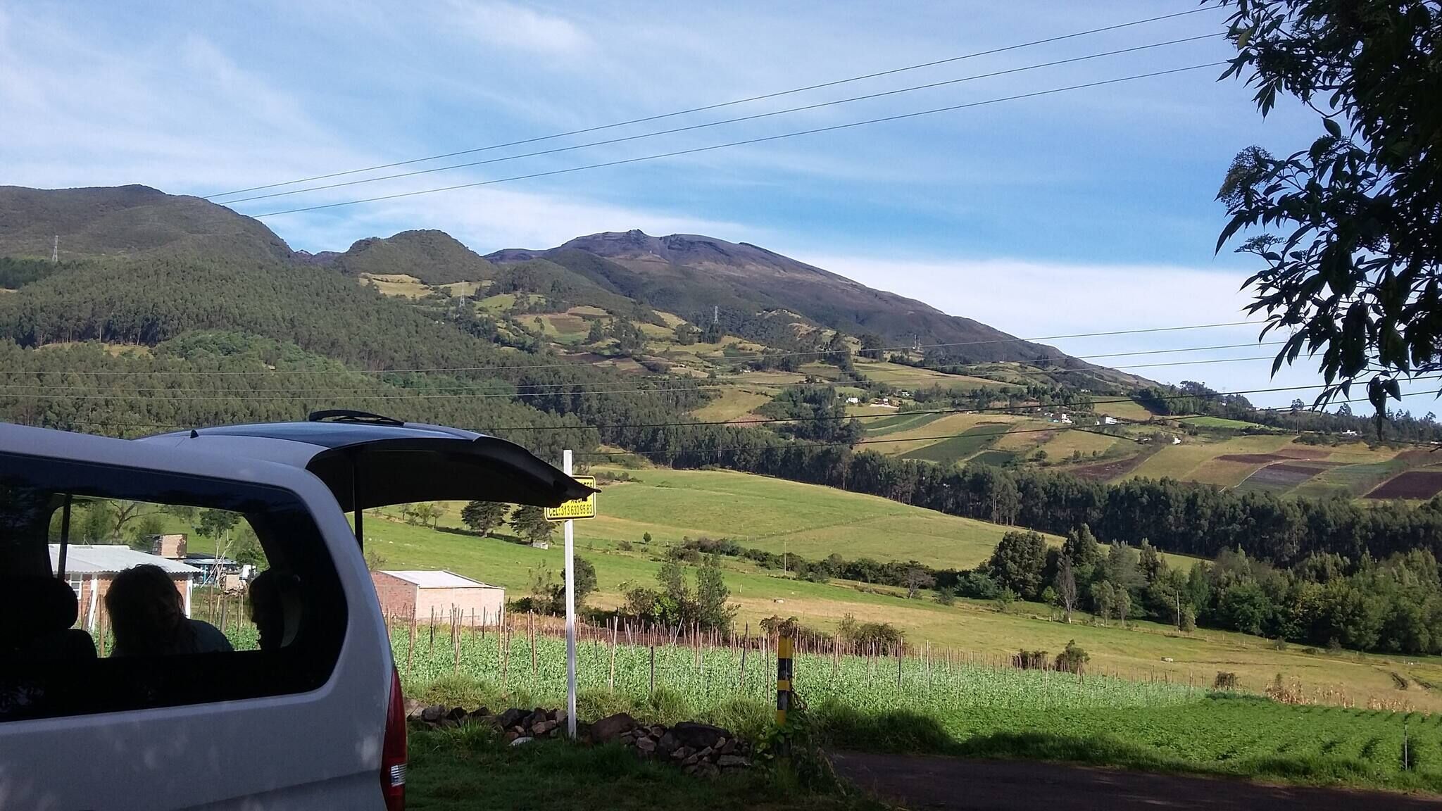 A white vehicle parked with its trunk open, overlooking a rural landscape with rolling hills, fields, and a mountain in the distance.