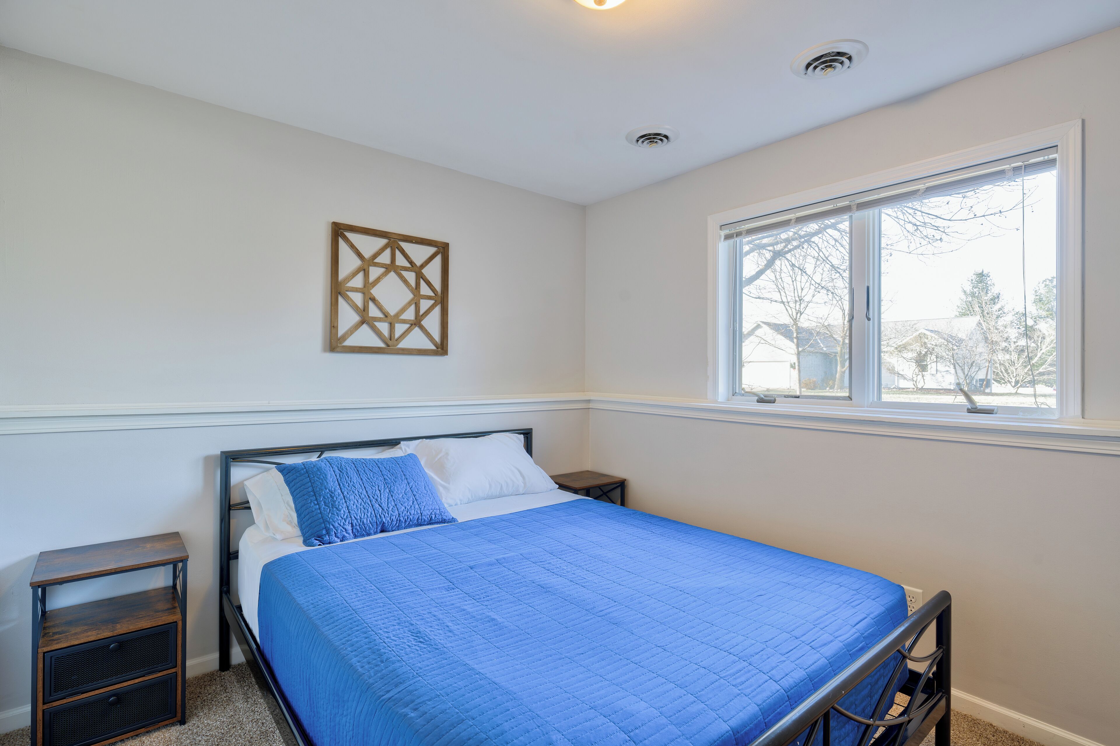 A bedroom with a bed, a nightstand, a window, and a decorative wall piece.