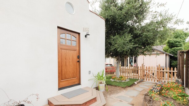 Una casa blanca con puerta de madera, buzón y un pequeño jardín con flores y cerca de madera.