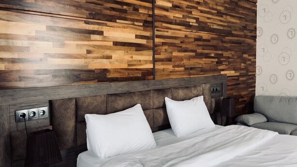 A hotel room with a wooden headboard, a bed with white bedding, a grey armchair, and a wall with a circular pattern. - Ire Palace (Khulo)