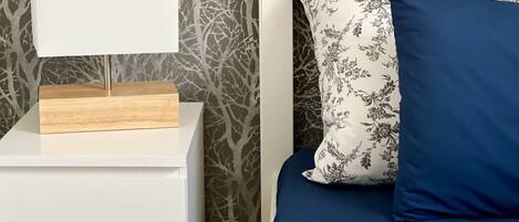 Una cama con una colcha de estampado floral y dos almohadas, una con motivos florales y otra azul lisa, frente a una pared con diseño de rama de árbol.
