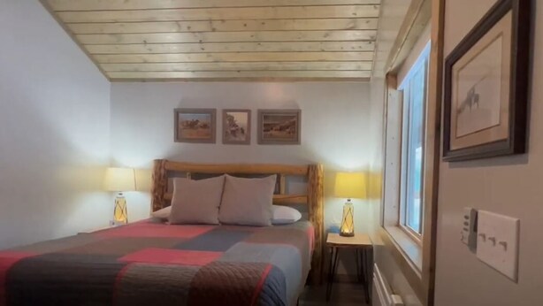 A bedroom with a wooden ceiling, a bed with a red and grey blanket, two bedside lamps, and three framed pictures on the wall. - King Room at Ponds Lodge Cabin 6 Unit 3 (Island Park)
