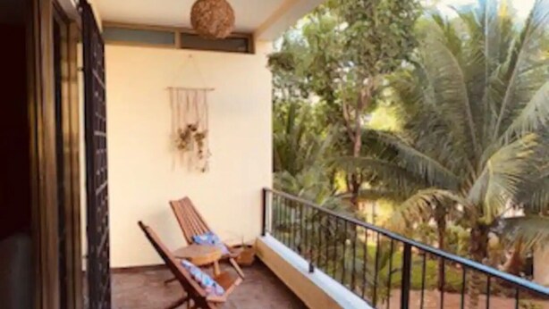 Un balcon avec une chaise en bois et une petite table, un luminaire suspendu, et une vue sur des palmiers et une végétation luxuriante.