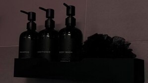Een zwarte plank met drie flessen, voorzien van de labels 'Shampoo', 'Conditioner' en 'Body Wash'.