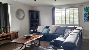 A living room with a sectional sofa, a wooden coffee table, a dark cabinet, and a large window with a view of trees.