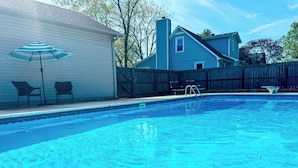 A swimming pool with a wooden deck, a covered patio area with chairs, and a house in the background.