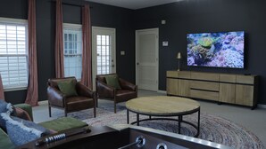 A living room with a pool table, leather sofas, a round coffee table, and a large flat-screen TV mounted on the wall.