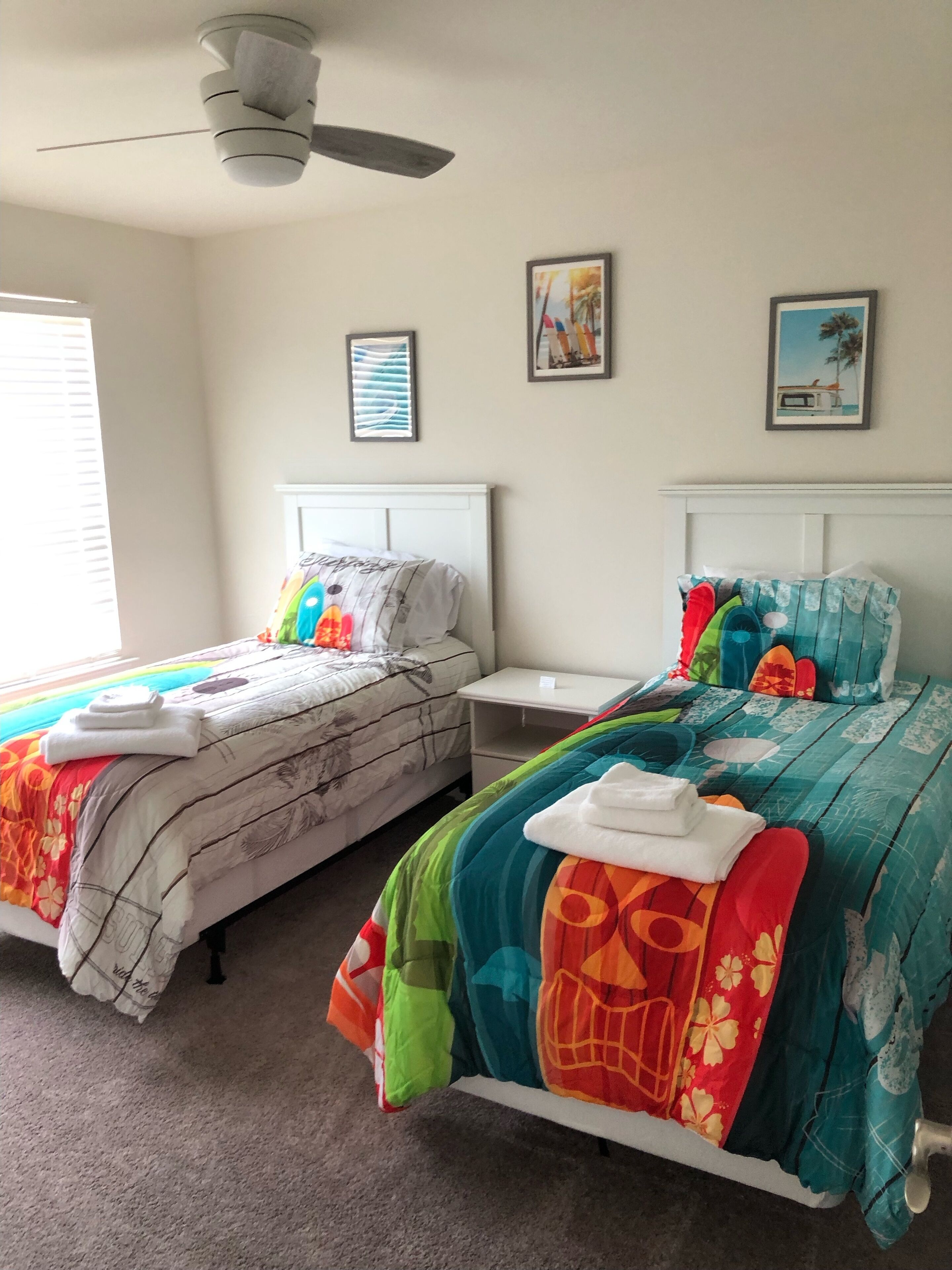 Una habitación con dos camas, cada una con ropa de cama y almohadas de colores, una mesita de noche blanca, un ventilador de techo y dos cuadros en la pared.