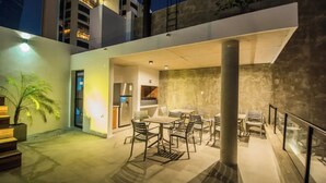 Un área de comedor exterior con diseño moderno, que incluye una mesa grande, sillas, un refrigerador y una planta en maceta.
