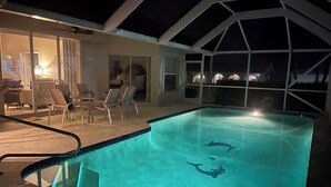 Une piscine avec deux dauphins, un patio couvert avec des chaises, et une vue sur le ciel nocturne à travers de grandes fenêtres.