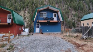 A two-story building with a balcony, a gravel driveway, and a forested hillside in the background. - Blue Cabin in Idaho Springs (Idaho Springs)