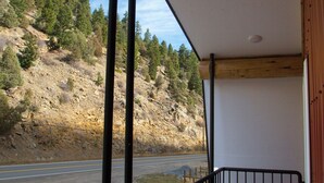 A wooden deck with a black railing, overlooking a road and a hillside with trees. - Blue Cabin in Idaho Springs (Idaho Springs)
