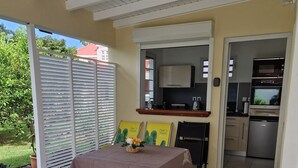 Un patio couvert avec une table dressée pour les repas, un coin cuisine équipé d’appareils électroménagers et une vue sur la verdure environnante.