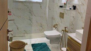 Ein modernes Badezimmer mit Marmorwänden, Toilette, Bidet, Waschbecken und Dusche. Auf dem Boden liegen grüne Handtücher und ein Korbwäscheschrank.