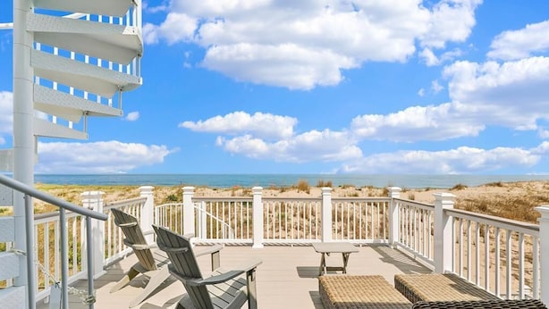 Eine Terrasse mit Stühlen und Tisch, mit Blick auf einen Strand mit Dünen und dem Meer.