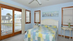 Une chambre à coucher avec un lit, un ventilateur de plafond, une fenêtre avec des stores, une table de chevet et un tableau au mur.