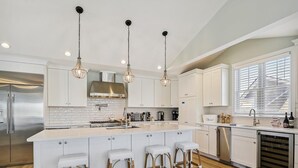 Una cucina moderna con mobili bianchi, un'isola centrale con sgabelli, elettrodomestici in acciaio inossidabile e illuminazione a sospensione.