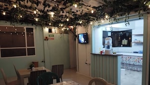 Un coin repas avec une table recouverte d’une nappe à motifs floraux, des chaises, un téléviseur accroché au mur et un espace cuisine visible à travers une porte ouverte.