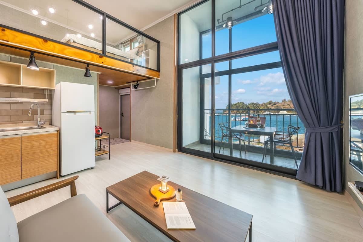 Una cocina moderna con horno empotrado, refrigerador y un comedor con mesa y sillas. Un gran ventanal ofrece vista al mar.
