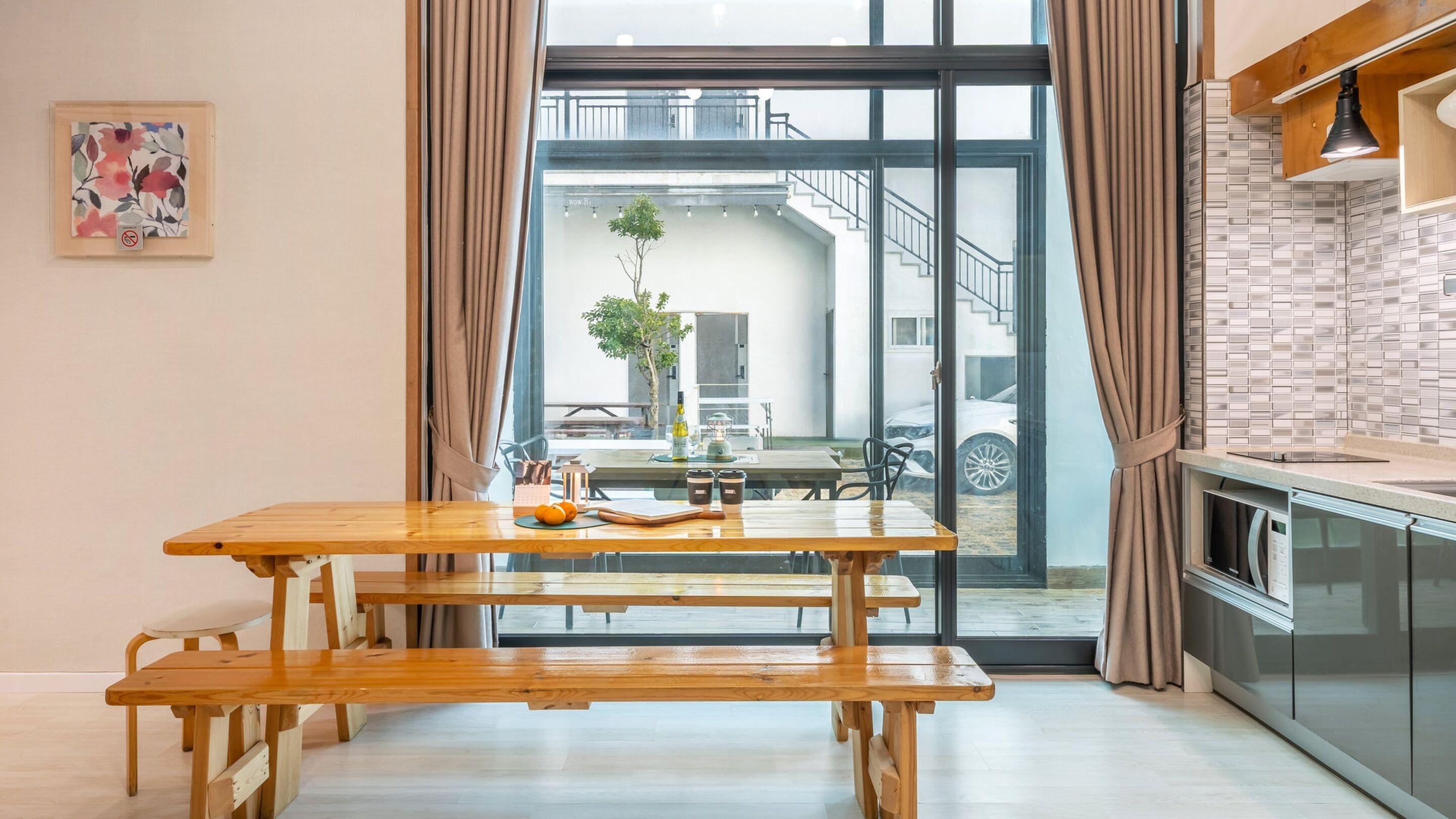 A modern kitchen with a wooden table and benches, a view of the outside, and a dining area with a plant and tableware.