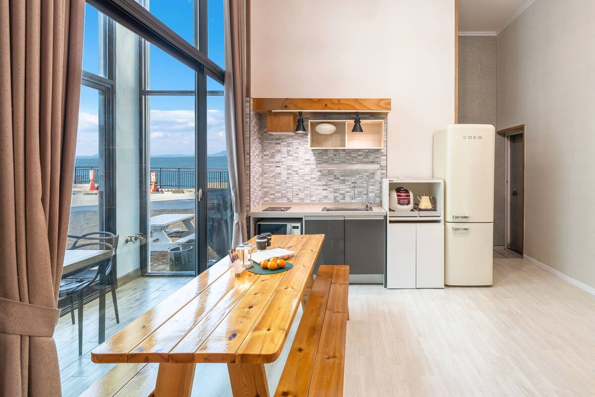 Cocina compacta con una mesa de madera, un refrigerador y vista al mar a través de amplios ventanales.