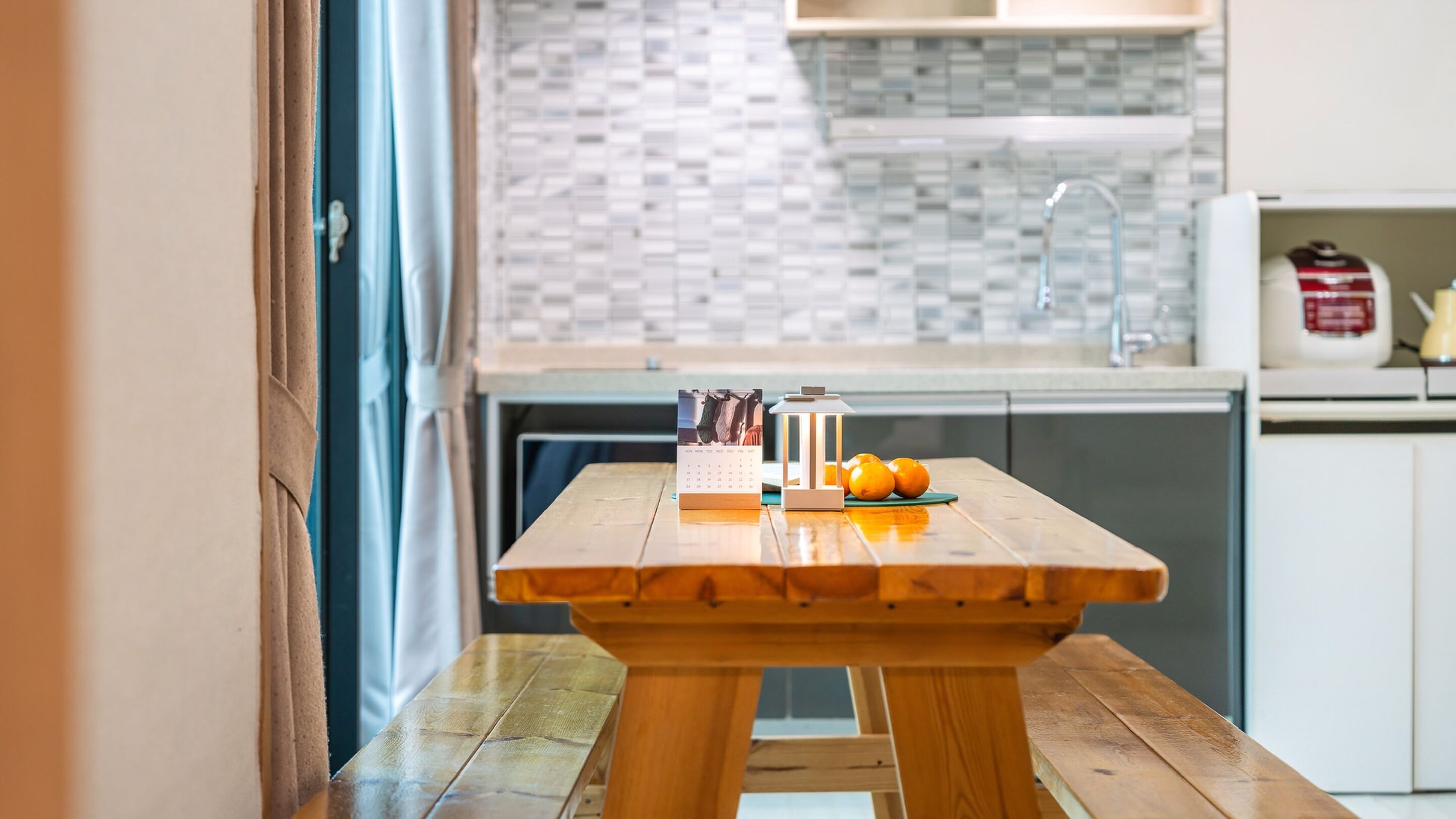 A wooden table with a calendar and oranges in a modern kitchen with a tiled backsplash.