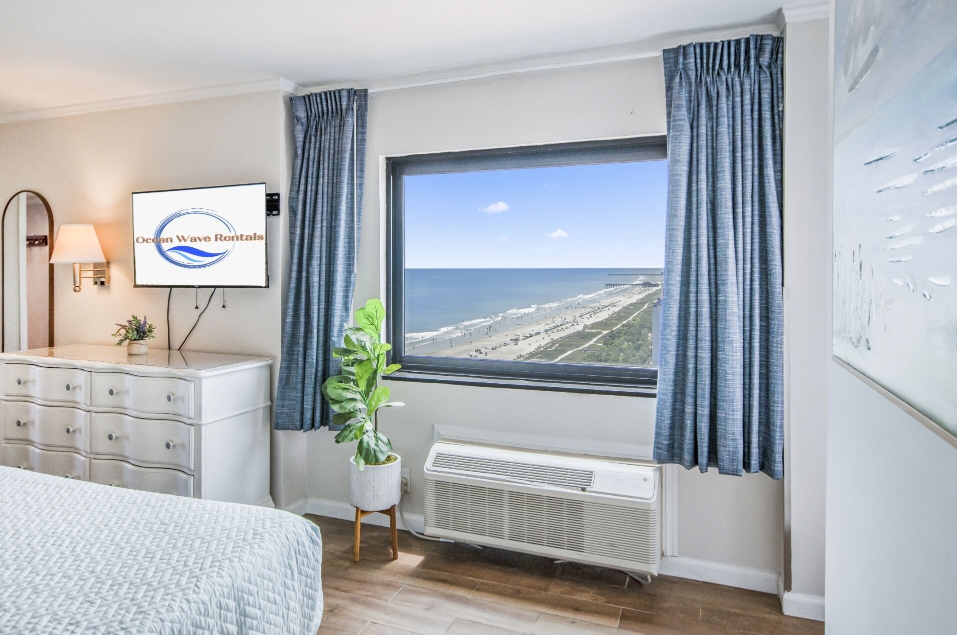 Quarto com cama, cômoda, planta em vaso, televisão presa à parede e janela com vista para a praia.