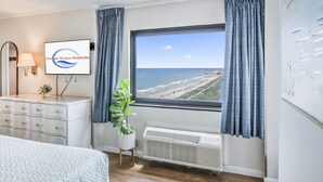 A room with a bed, a dresser, a potted plant, a wall-mounted TV, and a window with a view of the beach. - Corner Unit on the 16th Floor-Sweeping View, Ocean Front Balcony, New Paint (Myrtle Beach)