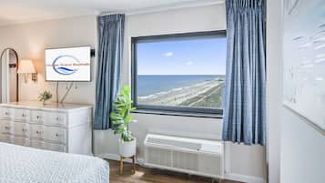 Quarto com cama, cômoda, planta em vaso, televisão presa à parede e janela com vista para a praia.