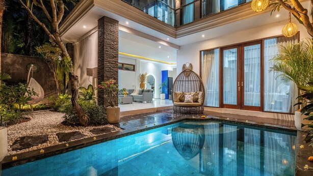 A modern indoor pool area with a clear pool, stone pathway, and a seating area with a wicker chair. - Villa Ashëla (By JB Villas) (Bali)