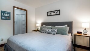 Un lit bien fait, recouvert d’une couverture grise, deux tables de chevet avec des lampes, un cadre décoratif au-dessus du lit et une porte sur le côté gauche.