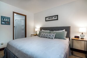 A neatly made bed with a grey quilt, two bedside tables with lamps, a framed sign above the bed, and a door to the left. - New! Close to Whitefish Lake, community hot tub & pool (Whitefish)