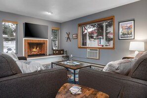 A living room with a fireplace, a television, and a large window with a view of trees and a building. - New! Close to Whitefish Lake, community hot tub & pool (Whitefish)