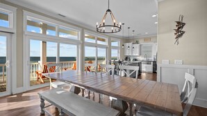 A spacious dining area with a large wooden table and benches, a chandelier, and a view of the ocean through large windows. - Hailo: Spacious 7-Bedroom Oceanfront Retreat in Oak Island, NC, perfect for up to 18 guests. (Oak Island)