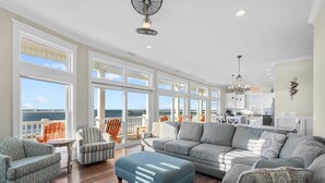 A spacious living room with a large gray sectional sofa, a blue ottoman, and a gray armchair. There are multiple windows offering a view of the ocean. - Hailo: Spacious 7-Bedroom Oceanfront Retreat in Oak Island, NC, perfect for up to 18 guests. (Oak Island)