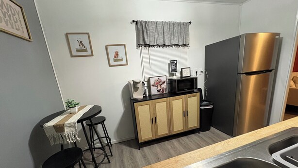 Een kleine keuken met een koelkast van roestvrij staal, een koffiezetapparaat en een magnetron op een houten aanrecht. Twee barkrukken en een tafeltje met een plantje.