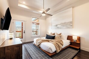 A bedroom with a bed, bedside table, lamp, ceiling fan, artwork, and a door with a view.