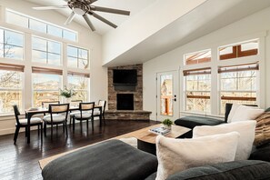 A spacious living room with a fireplace, a dining area with chairs, and large windows offering views of the outdoors.
