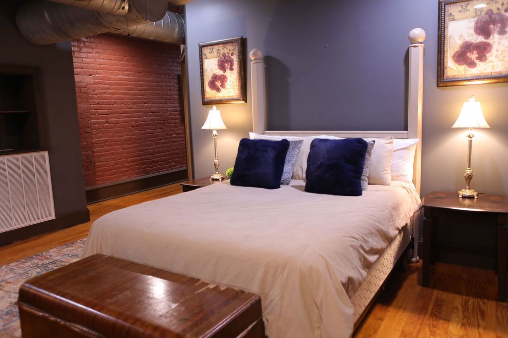A neatly arranged bedroom with a large bed, two bedside lamps, a wooden nightstand, and a brick accent wall.