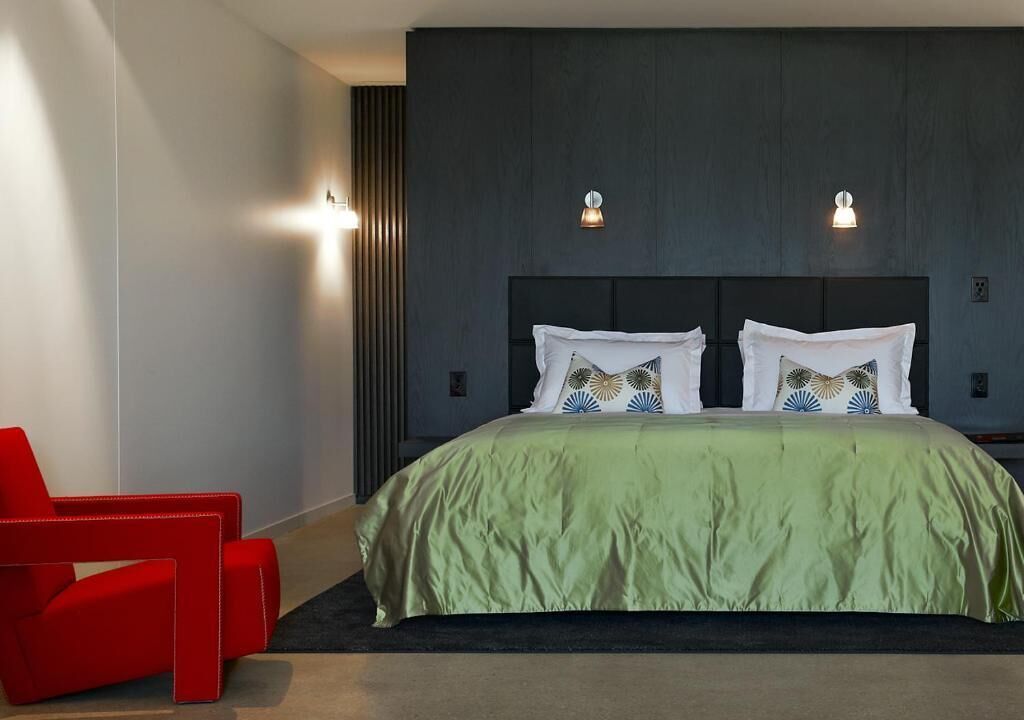 Ein modernes Hotelzimmer mit einem großen Bett, zwei Wandleuchten, einem roten Sessel und einem dunklen Holz-Kopfteil.