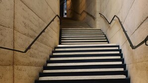 Una escalera con escalones negros y barandillas de metal, que asciende dentro de un edificio de paredes de piedra.